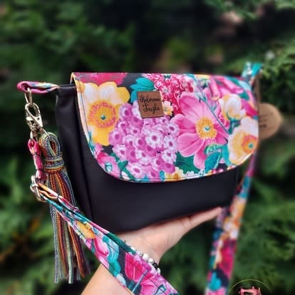 Hand holding a crossbody bag with floral fabric top and black leather bottom, featuring a colorful strap and tassel charm
