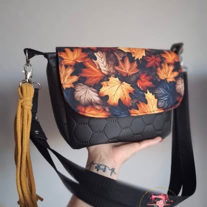 Person holding a black quilted shoulder bag with autumn leaf pattern, featuring a golden tassel charm and adjustable strap