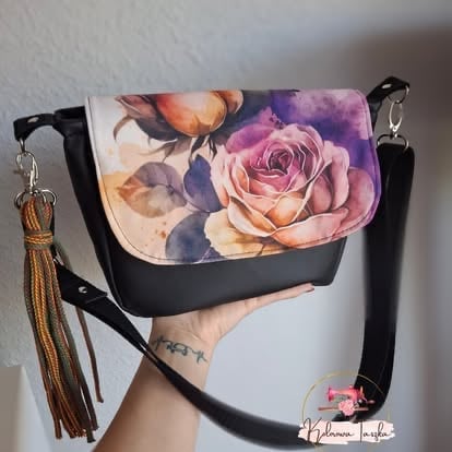Black crossbody bag with watercolor floral rose design on front flap and tan tassel charm, held by person with visible tattoo