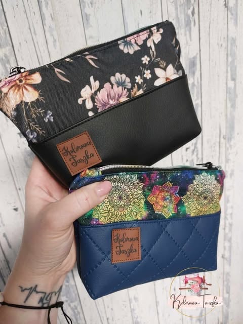 Hand holding two decorative pouches with floral and patterned fabric tops and solid fabric bottoms, featuring leather labels