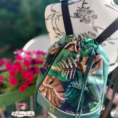 Colorful tropical leaf print drawstring backpack with black straps held up outdoors, blurred pink flowers in background