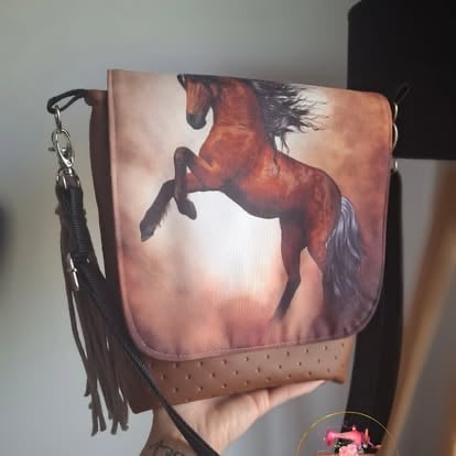 Brown leather shoulder bag featuring a rearing horse image with black fringe tassel, displayed against white wall